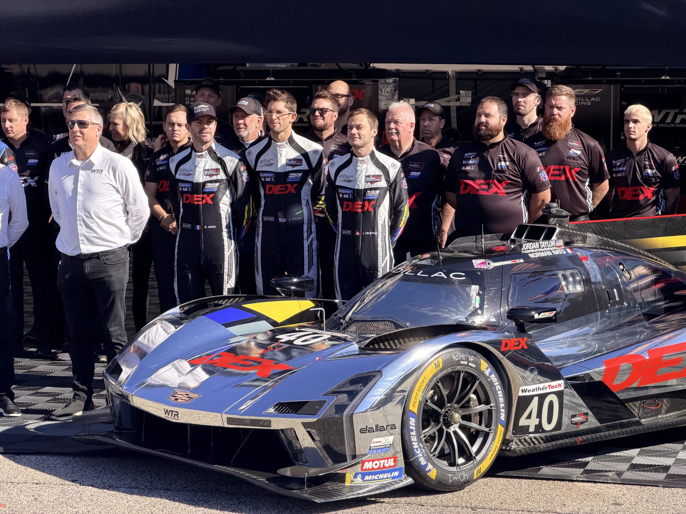 All the personnel of a Cadillac LMDh race team lined up behind the car facing right to left